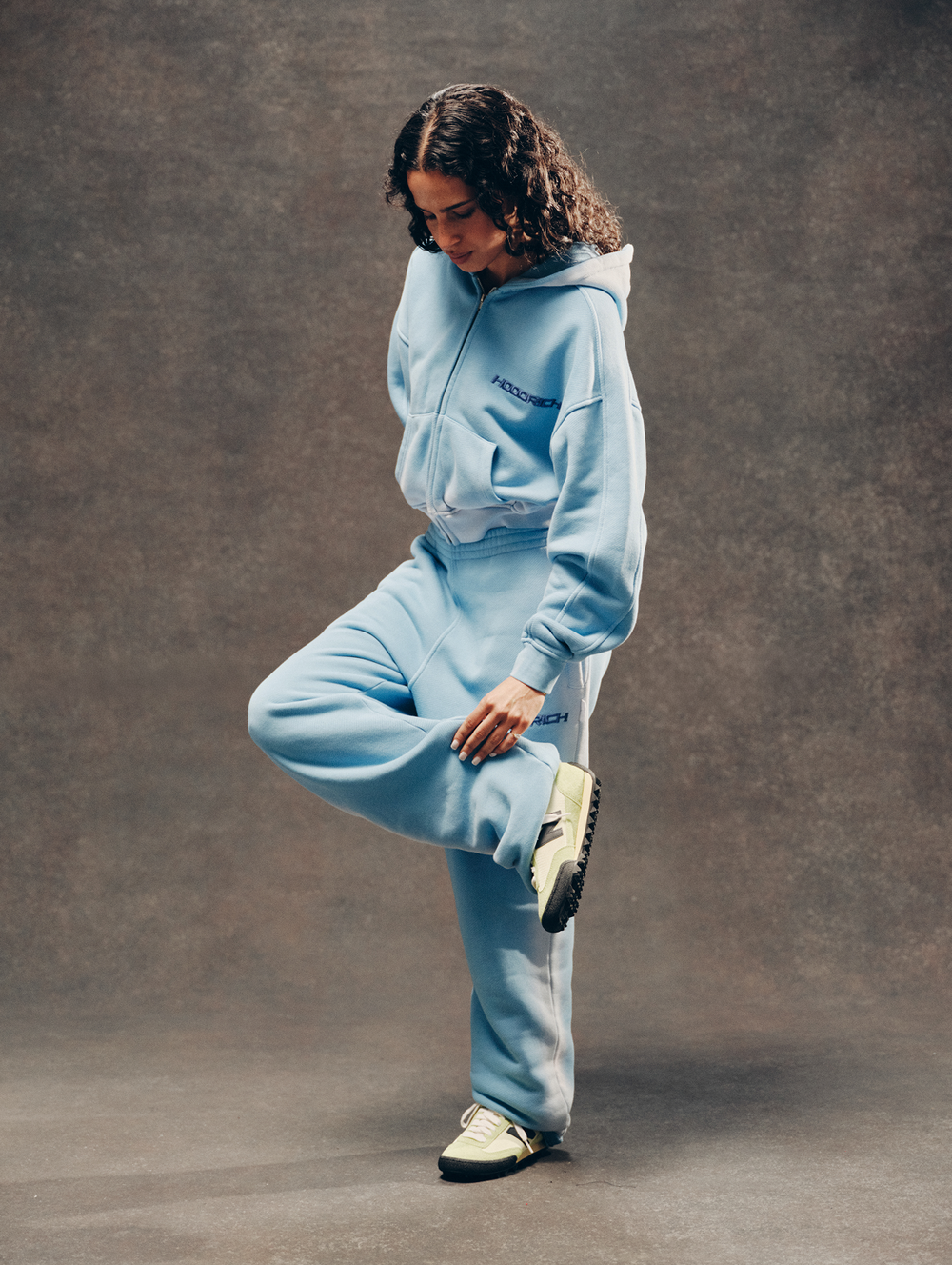 A model wearing a light blue tracksuit consisting of a zip-up hoodie and matching sweatpants, posing against a textured brown studio backdrop.