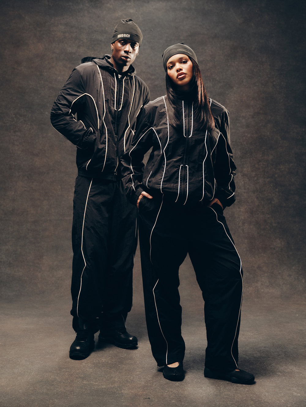 Two models wearing matching black streetwear tracksuits with white piping details, standing against a dark textured studio backdrop.