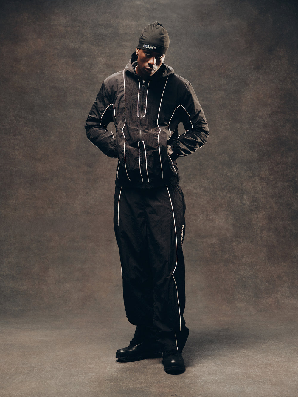 A model wearing a black hooded tracksuit with white contrast stitching, a black beanie with white text, and black boots, standing against a textured brown studio backdrop.