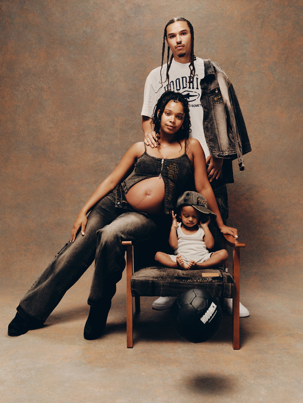 A family portrait of a pregnant woman, a man standing behind her, and a young child sitting on a wooden stool. The woman wears a dark brown cropped vest and wide-leg jeans, while the man wears a white graphic t-shirt and a dark denim jacket.