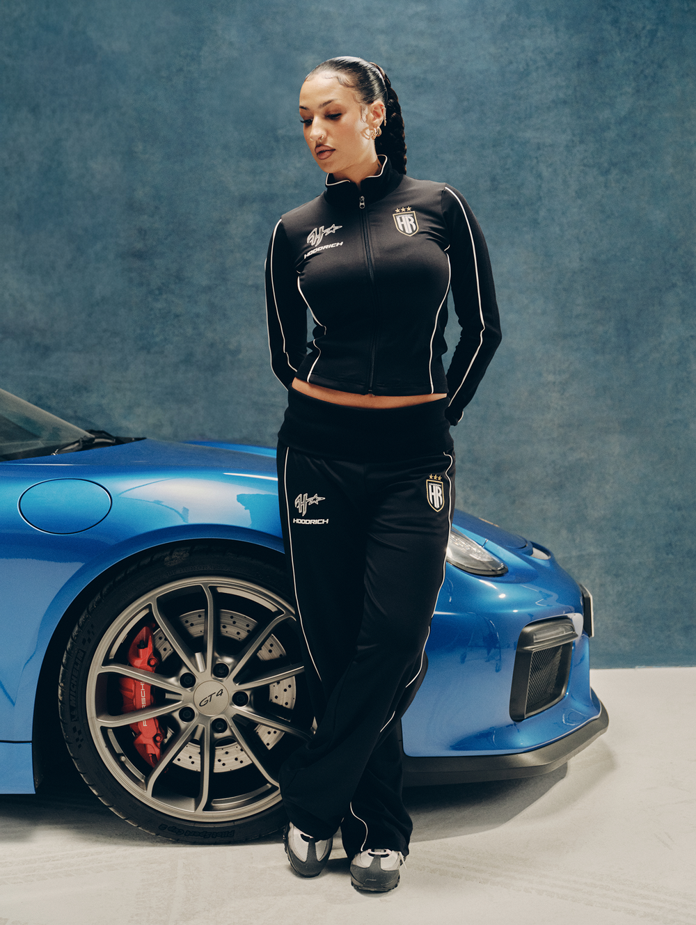 A woman modeling a black 'HOODRICH' tracksuit with white piping and logos, standing next to a Porsche sports car against a textured blue-gray wall.