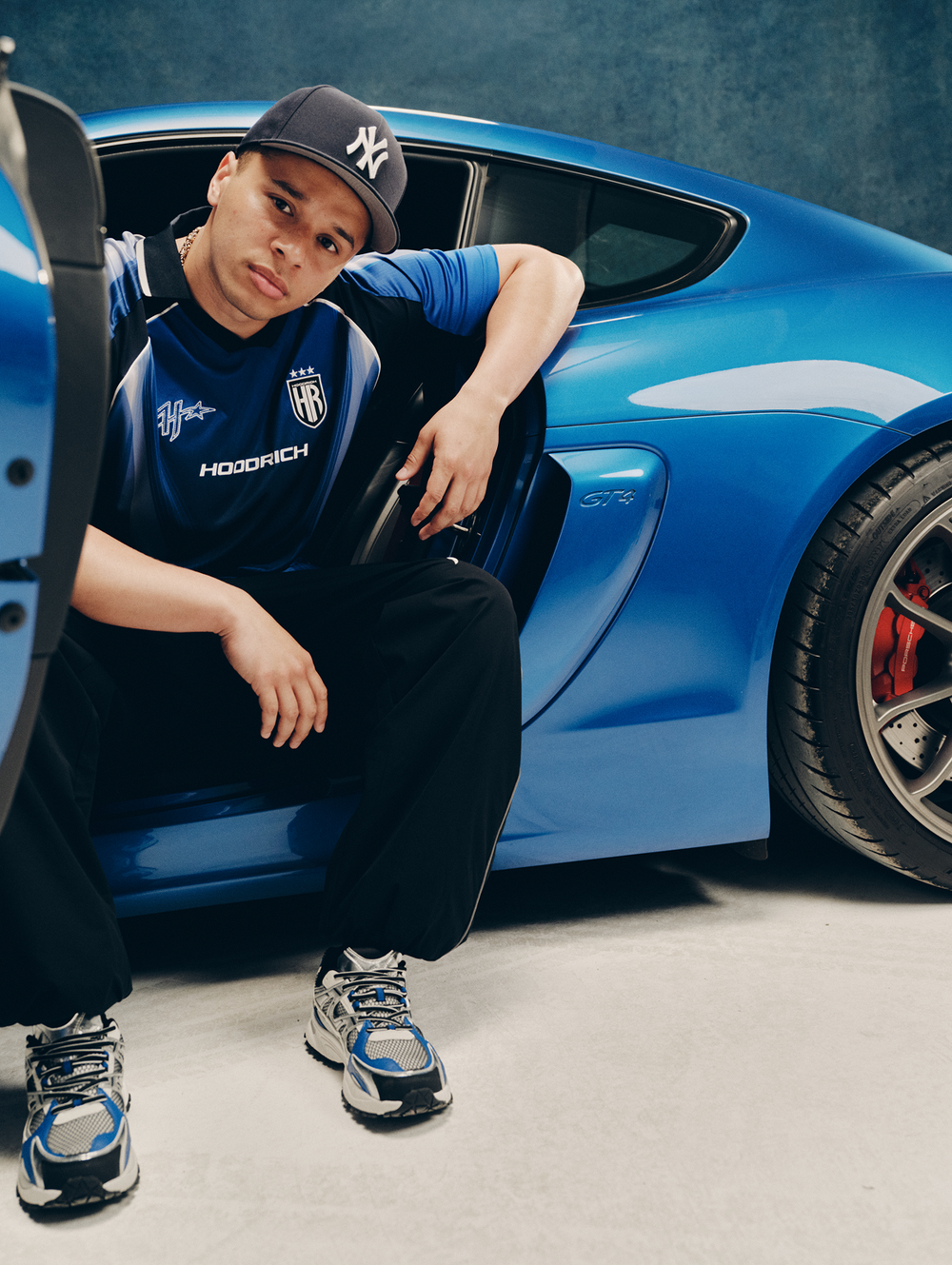 A man wearing a blue and black HOODRICH sports jersey, black pants, and a black New York Yankees cap leans against a bright blue Porsche sports car in a studio setting.