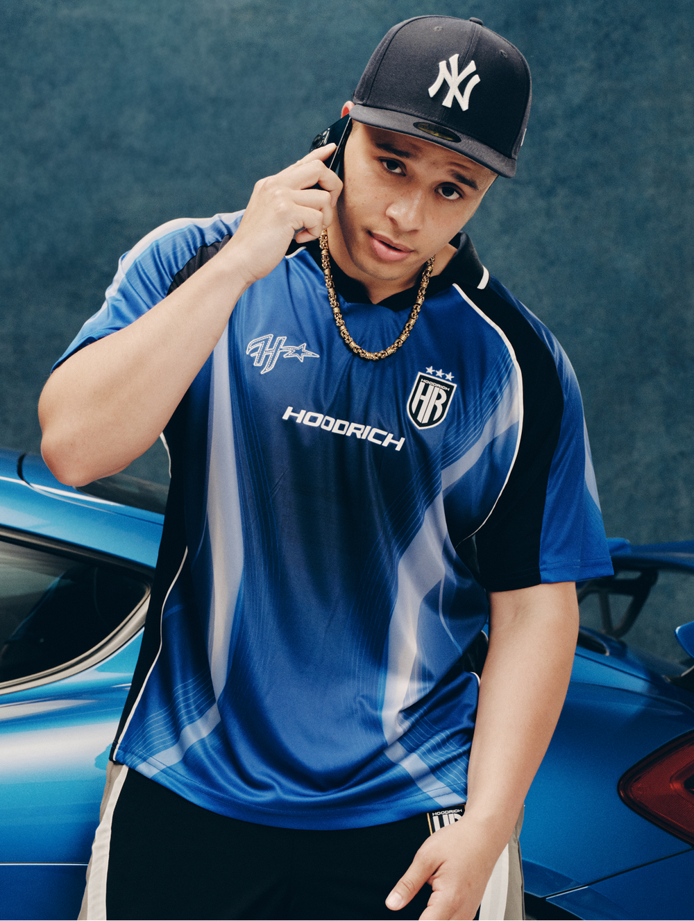 A male model wearing a blue 'HOODRICH' football jersey and a navy new york yankees cap. Staring into the camera holding his phone to his ear. Leaning against a blue Porsche sports car.