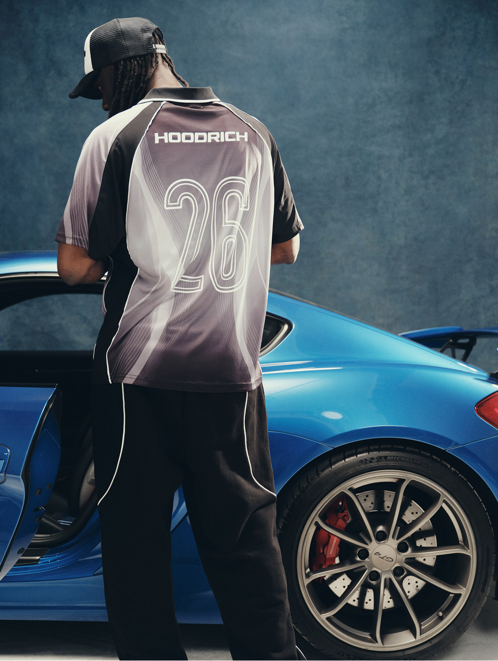 A man stands beside a bright blue Porsche sports car, shown from the back wearing a dark gray and black raglan-style jersey with 'HOODRICH' and the number '26' printed on the back, along with black pants and a black cap.