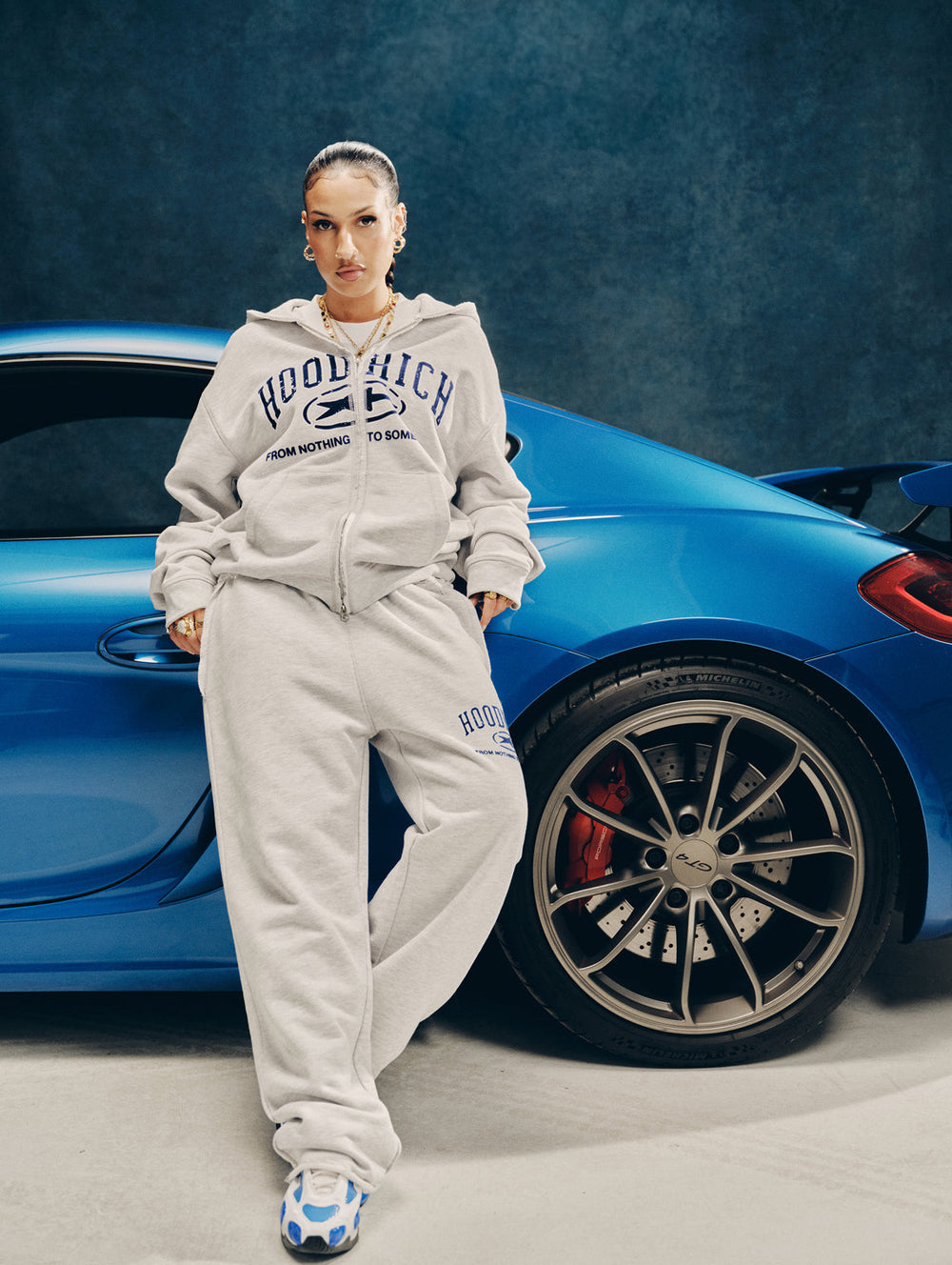 A female model wearing a light gray 'HOODRICH' branded zip-up hoodie and matching sweatpants stands next to a bright blue Porsche sports car against a dark textured studio backdrop.