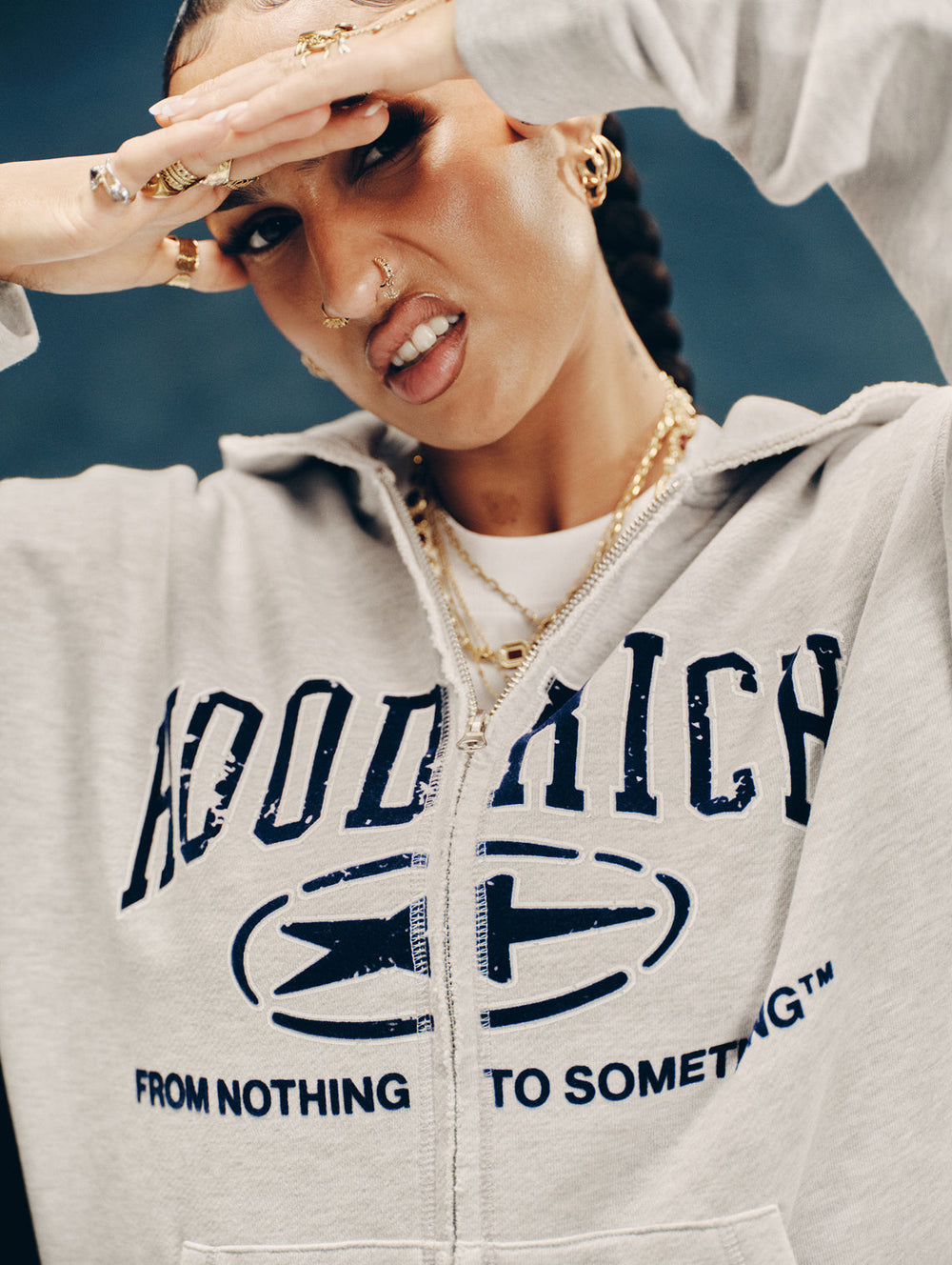 A close-up lifestyle shot of a female model wearing a light gray zip-up hoodie with a navy blue graphic print that reads 'HOOD RICH' and 'FROM NOTHING TO SOMETHING'. The model is also wearing gold jewelry, including a layered chain necklace and rings.