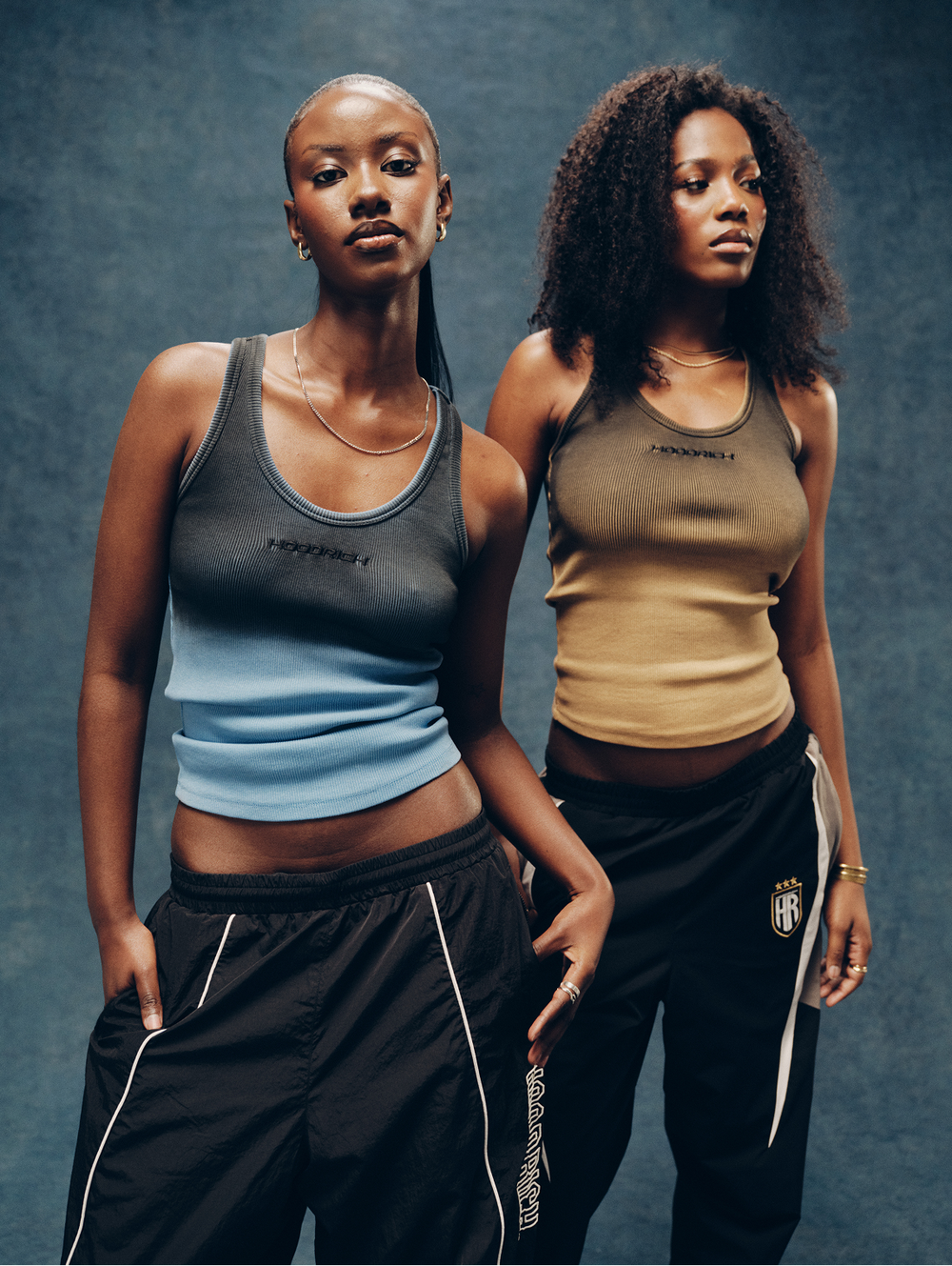 Two models posing in matching athletic wear from the brand 'HOODRICH', featuring a dark gray and light blue tank top on the left, and a beige and tan tank top on the right, paired with black track pants with white piping.