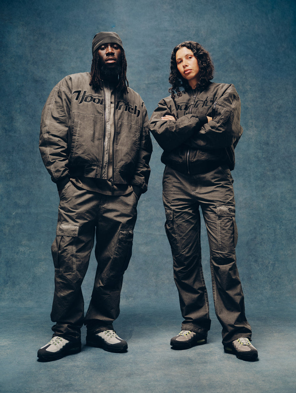 Two models wearing matching dark gray oversized tracksuits with 'HOODRICH' text on the chest, standing against a textured blue studio backdrop.