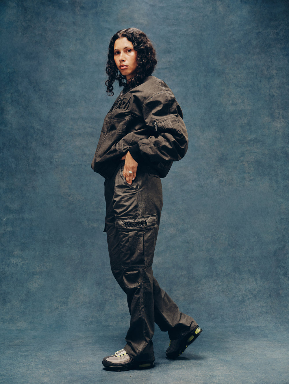A female model stands in profile wearing a dark oversized 'HOODRICH' utility jacket and matching cargo pants against a textured blue studio backdrop.