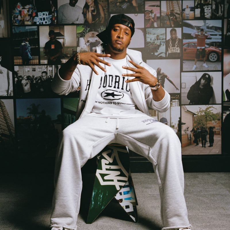 A man wearing a white HOODRICH sweatshirt and matching sweatpants sits on a green and white chair made with skateboards in a studio setting with a collage wall behind him. The HOODRICH brand logo is visible on the sweatshirt and bottoms.