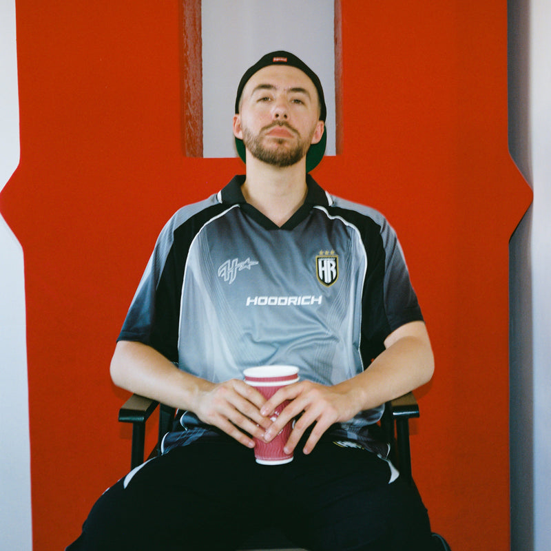 A man wearing a black and gray soccer jersey with 'HOODRICH' printed on the front, sitting in a chair and holding a pink cup in front of a bold red and white wall.
