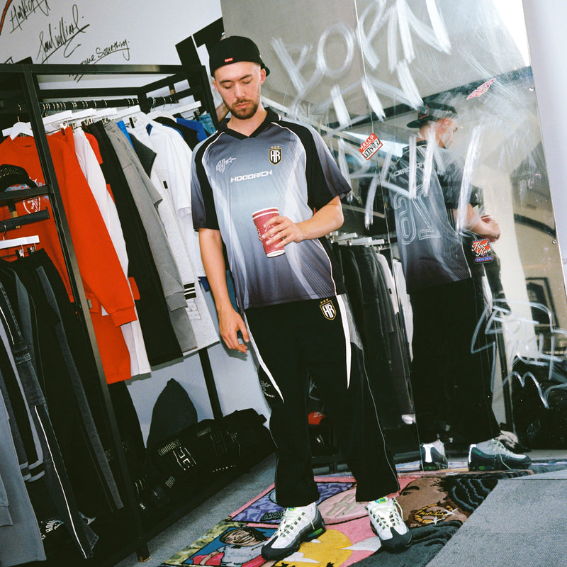 A man wearing a black and gray HOODRICH soccer jersey, black track pants, and white sneakers stands in a retail store holding a red cup. The store has clothing racks, graffiti on the glass, and a mirror reflecting the scene.